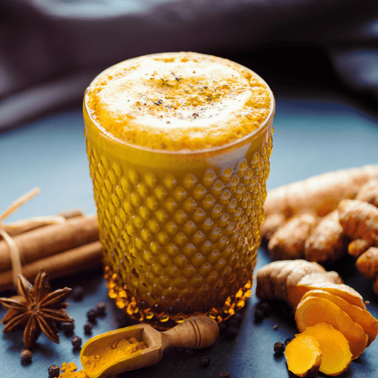 Verre de lait d'or entouré de curcuma frais, d'épices en poudre (cannelle, poivre noir), et de bâtons de cannelle, illustrant une boisson ayurvédique riche en bienfaits pour la santé.