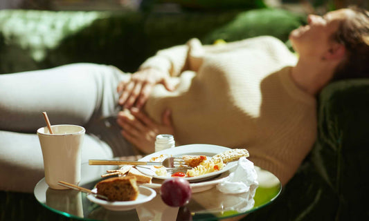 Personne souffrant de ballonnements, assise sur un canapé avec un repas léger, illustrant les inconforts digestifs et l'importance des solutions naturelles comme le charbon végétal ou les graines de fenouil.