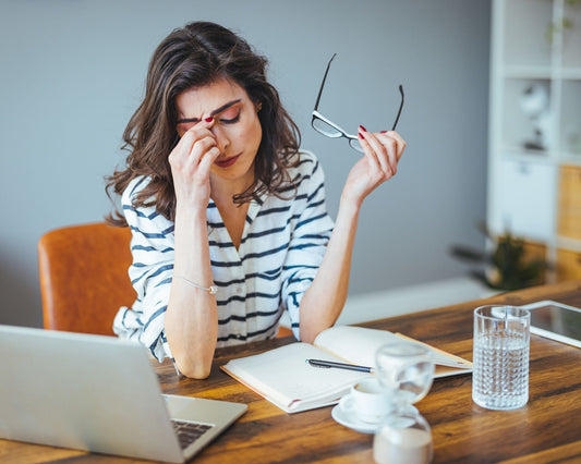 Femme assise à son bureau, les yeux fermés, se tenant le front et les lunettes, manifestant des signes de fatigue, de stress et de surcharge mentale, devant un laptop, un agenda et un stylo.