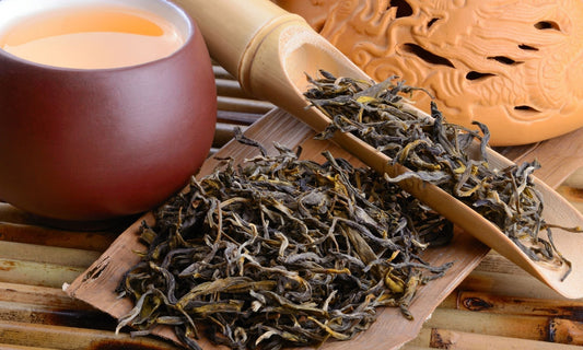 Feuilles de thé séchées dans une cuillère en bambou, à côté d'une tasse de thé infusé dans une théière en terre cuite