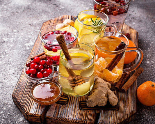 Sur une planche en bois, plusieurs tasses en verre remplies d’eau infusée avec des tranches de citron, de clémentine et de baies d’églantier, accompagnées de gingembre frais, de branches de romarin, de bâtons de cannelle et d’un pot de miel.