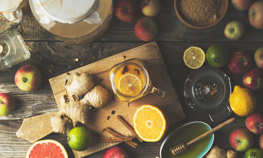 Vue du dessus d'ingrédients pour oxymel maison — pommes, citron, orange, pamplemousse, gingembre frais, bâtons de cannelle, miel et bocal en cours de macération sur une table en bois.