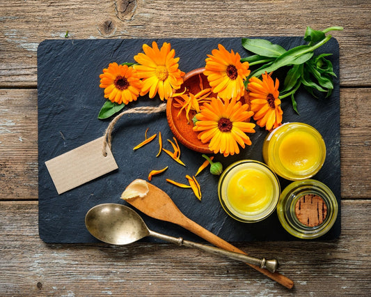Fleurs fraîches de calendula posées sur une ardoise avec une cuillère et une spatule en bois, accompagnées de pots de pommade au calendula maison