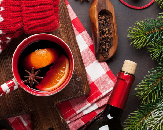 Tasse de vin chaud de Noël posée sur une planche en bois avec rondelles d’orange, anis étoilé, bouteille de vin rouge et branche de sapin dans une ambiance festive.