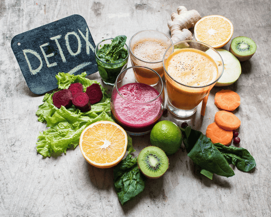 Ardoise avec l'inscription "DETOX" à côté de verres de jus frais, entourés de fruits et légumes colorés.