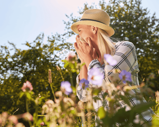 Femme portant un chapeau dans un champ fleuri, éternuant dans un mouchoir à cause des allergies printanières et de l’arrivée des pollens.