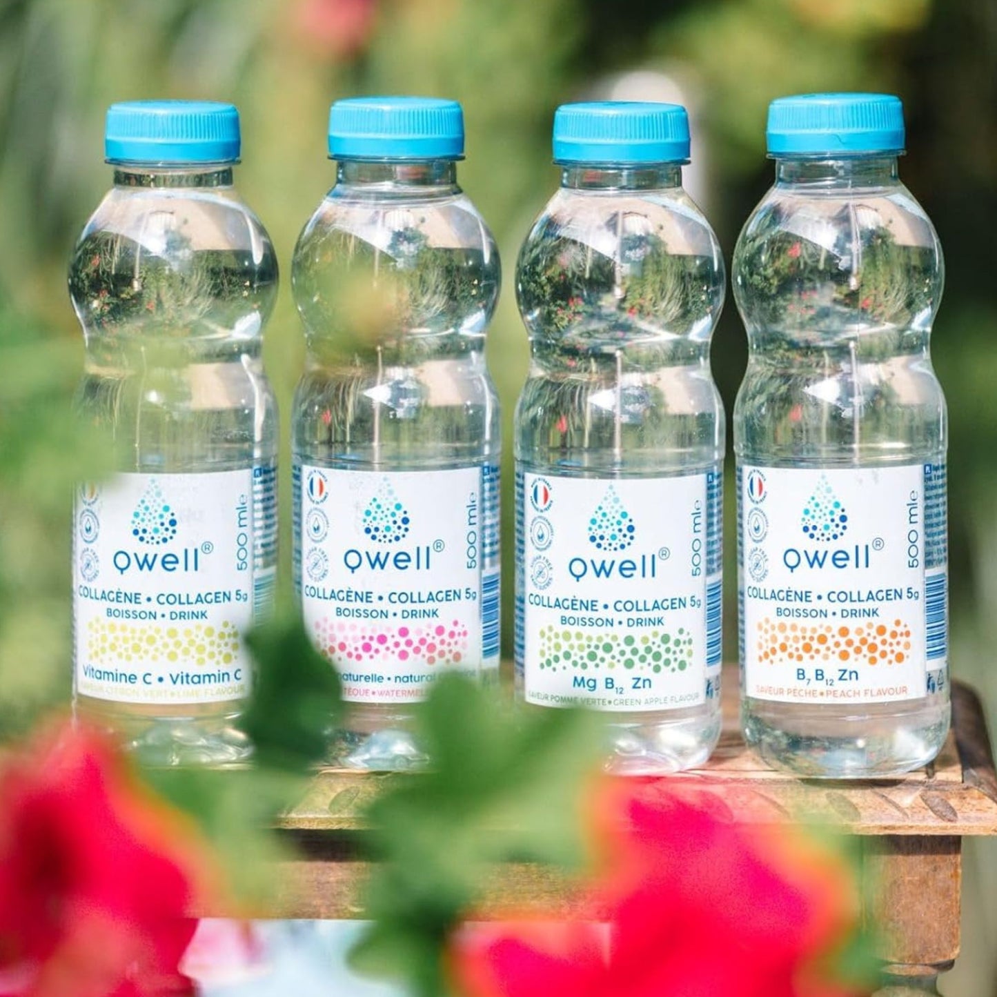 Les quatre bouteilles de boisson au collagène Qwell posées sur un banc de bois entouré de fleurs rouges — saveurs citron vert, pastèque, pomme verte et pêche, enrichies en vitamines et minéraux