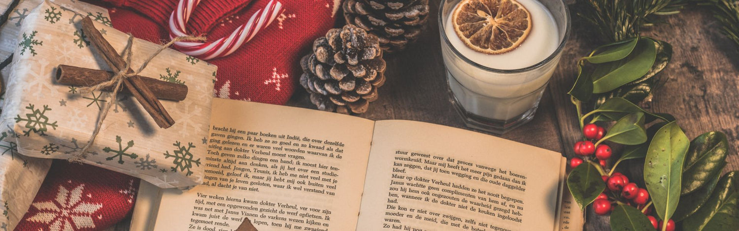 Livre ouvert sur une table en bois, à côté un pull de Noël, un cadeau emballé, des branches de cannelle, pommes de pin, branche de sapin et un verre de lait avec une tranche d’orange séchée, décor festif de Noël.