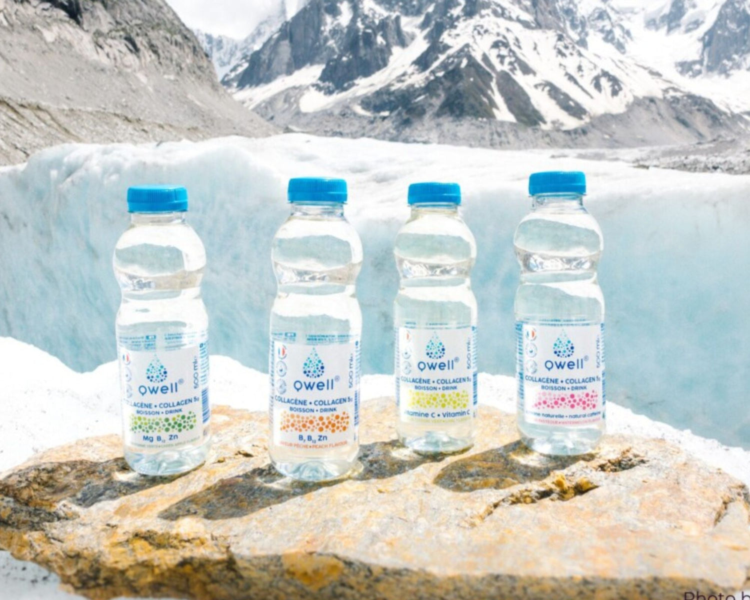 Quatre bouteilles Qwell d’eau au collagène posées sur une pierre au bord d’un lac avec des montagnes en arrière-plan, illustrant une hydratation naturelle et un mode de vie sain