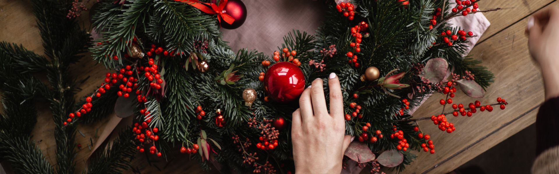 Mains en train de confectionner une couronne de l’Avent sur une table en bois, avec branches de sapin, boules de Noël rouges et fruits rouges, ambiance festive et artisanale.