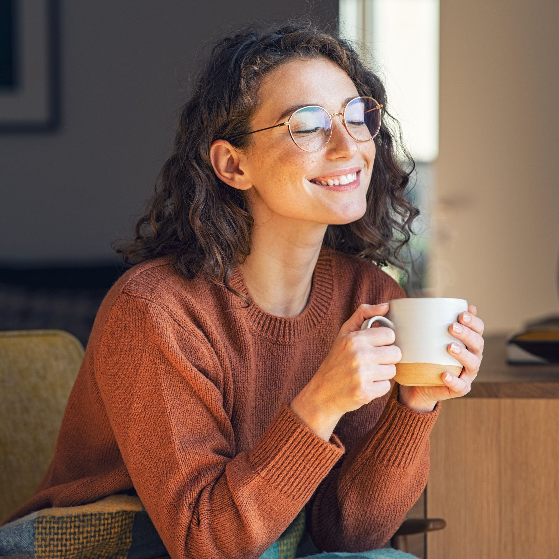 Femme aux cheveux bouclés et lunettes rondes, les yeux fermés, tenant une grande tasse de tisane entre ses mains, dans une atmosphère cosy et apaisante