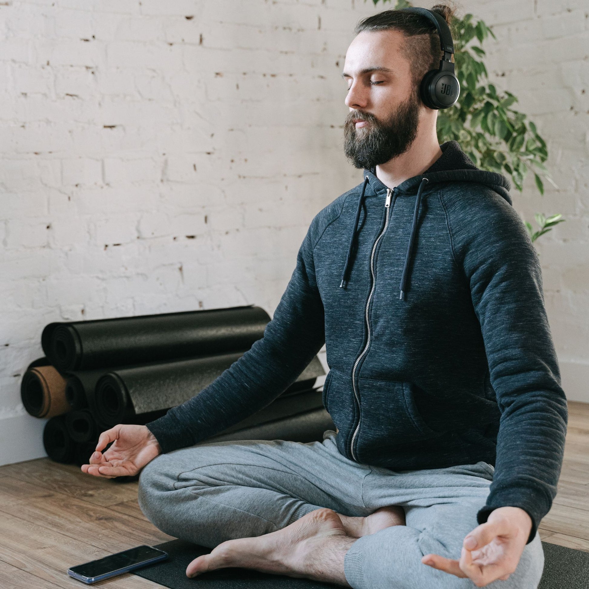 Homme barbu assis en position lotus sur un tapis de yoga, casque audio sur les oreilles, les yeux fermés, dans un espace calme et lumineux