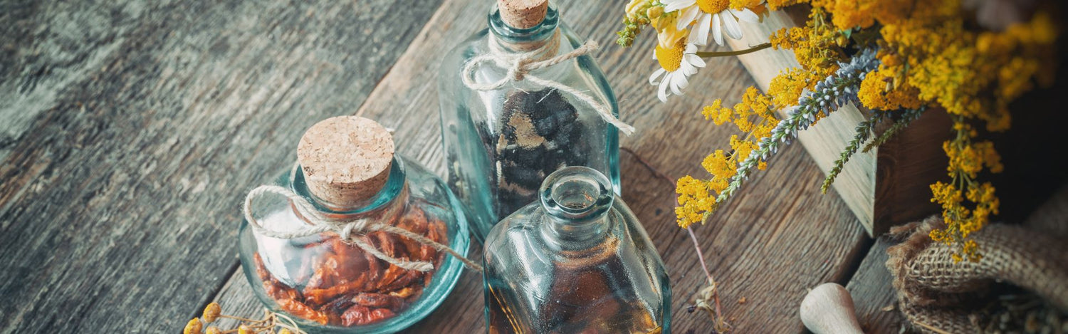 Trois récipients en verre remplis de remèdes naturels posés sur une vieille table en bois, accompagnés d’une boîte en bois contenant des plantes médicinales fraîches.