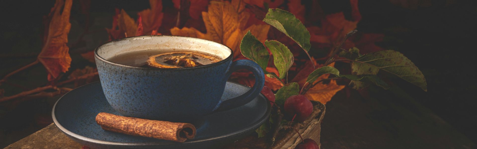 Tasse bleue de tisane avec un morceau d’orange séché et un bâton de cannelle sur un meuble en bois, entourée d’une branche aux fruits rouges et de feuilles aux couleurs d’automne